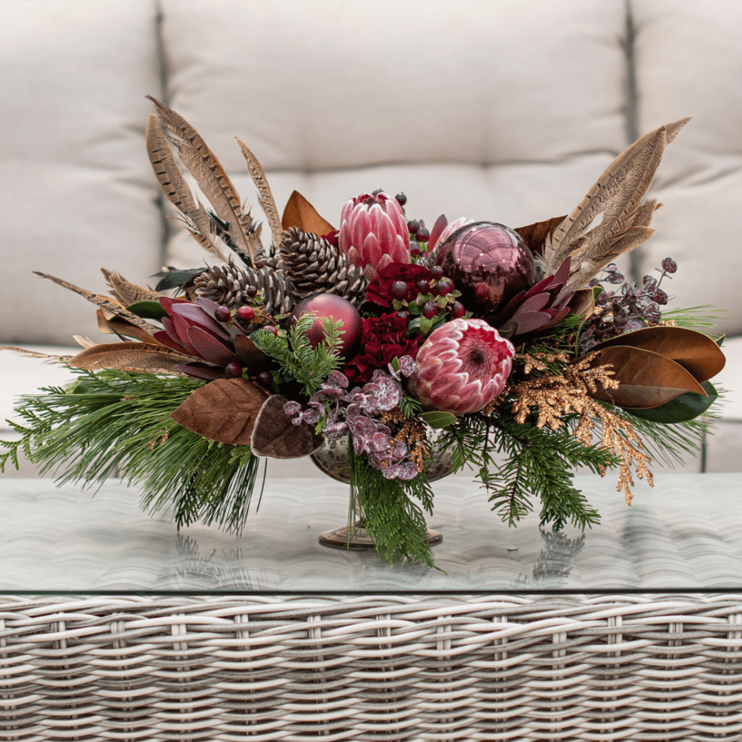 111 Festive floral arrangement with pink proteas, pinecones, feathers, and greenery on a wicker table.