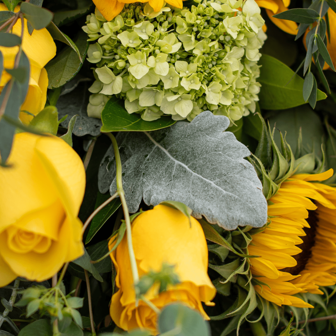 Vibrant bouquet of yellow roses, sunflowers, green hydrangeas, and foliage, creating a lush and cheerful floral arrangement.