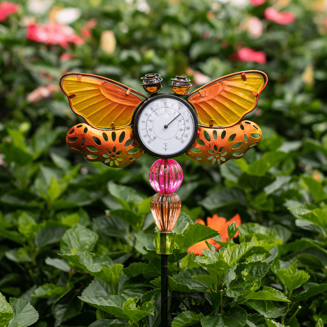 Colorful butterfly garden thermometer amidst lush green foliage, displaying temperature in Celsius and Fahrenheit.