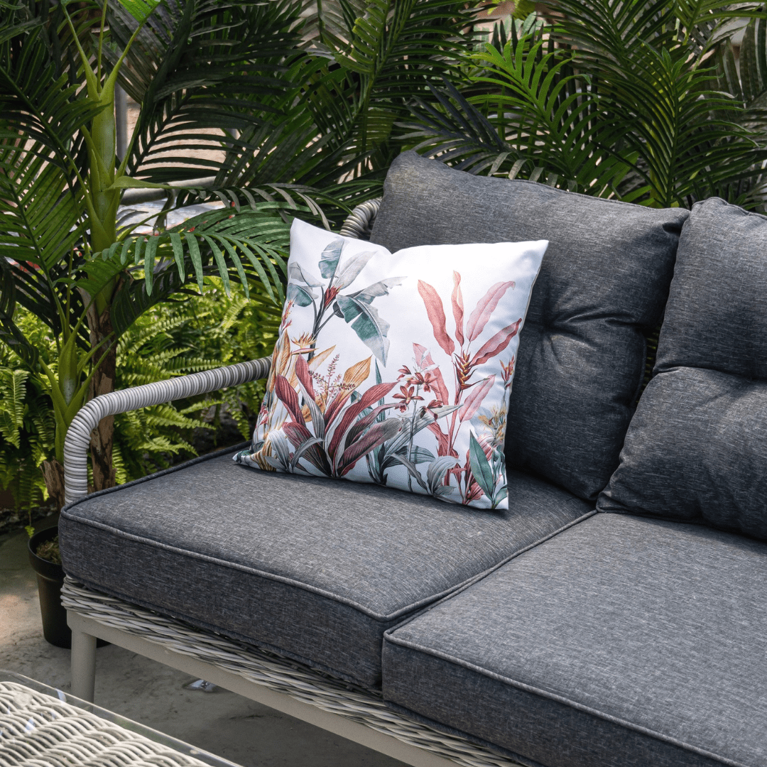 Outdoor patio with grey sofa and tropical-patterned cushion surrounded by lush green plants.