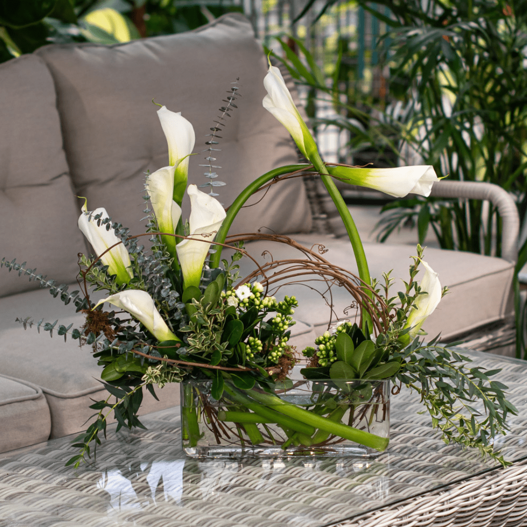 Elegant calla lily arrangement in a glass vase on a patio table, surrounded by greenery and wicker furniture.