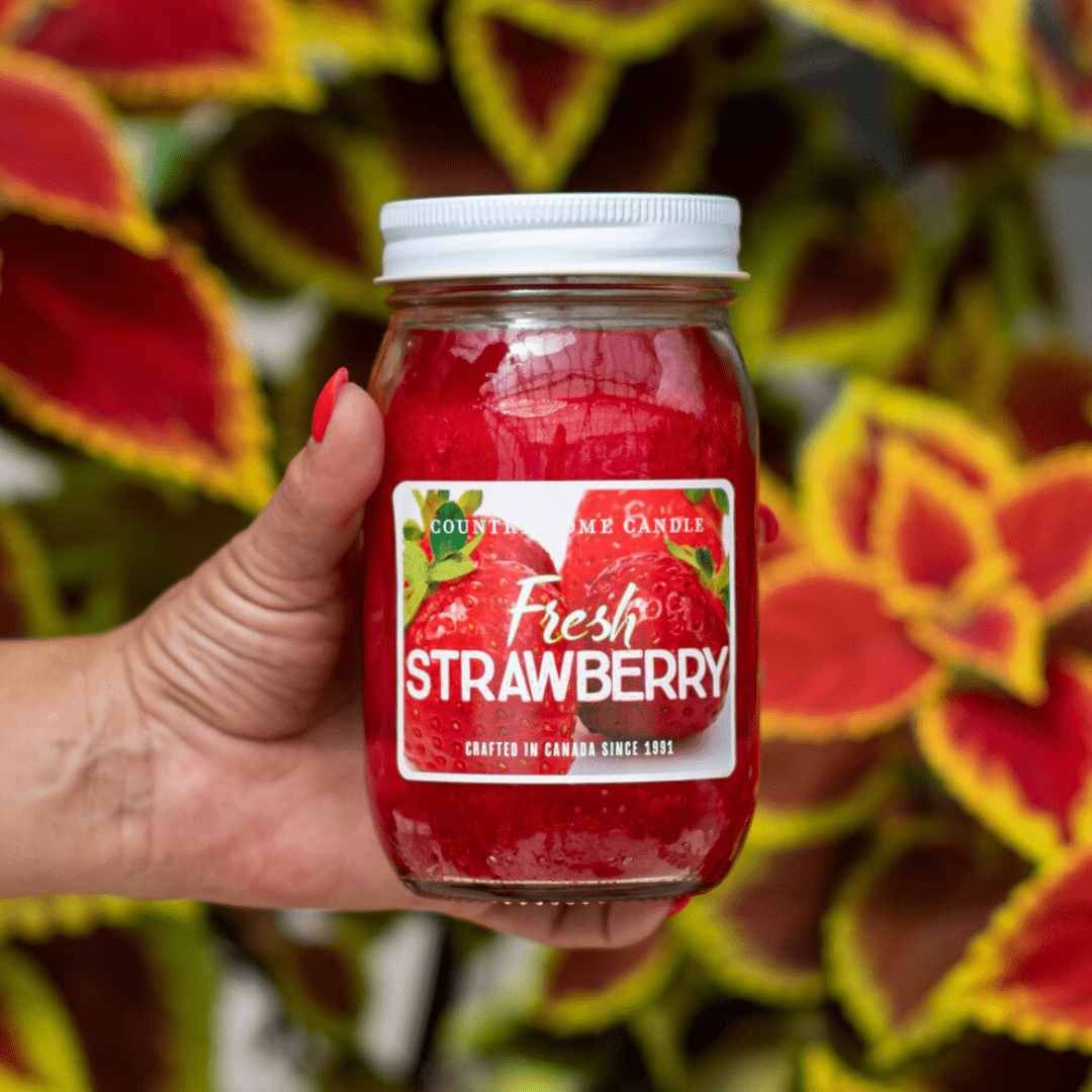 Hand holding a jar of Fresh Strawberry scented candle with vibrant leafy background. Perfect for a cozy ambiance.
