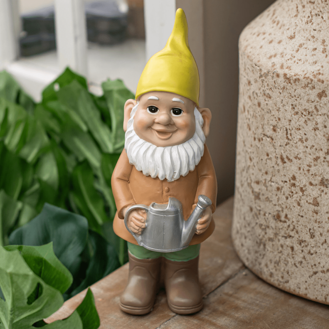 Cheerful garden gnome with yellow hat holding watering can beside leafy plants and stone urn.