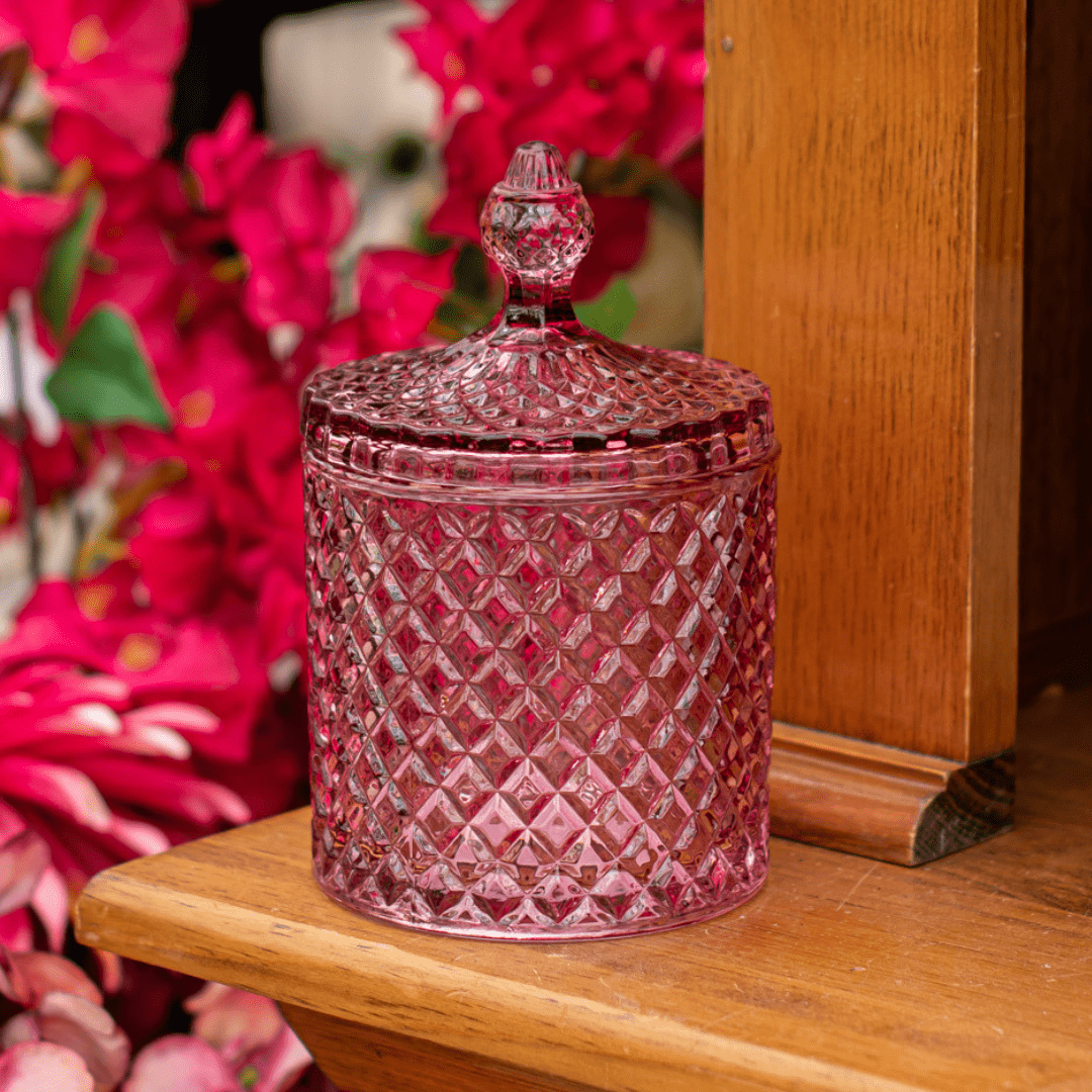 Ornate pink glass jar with a lid on a wooden shelf, surrounded by vibrant magenta flowers.