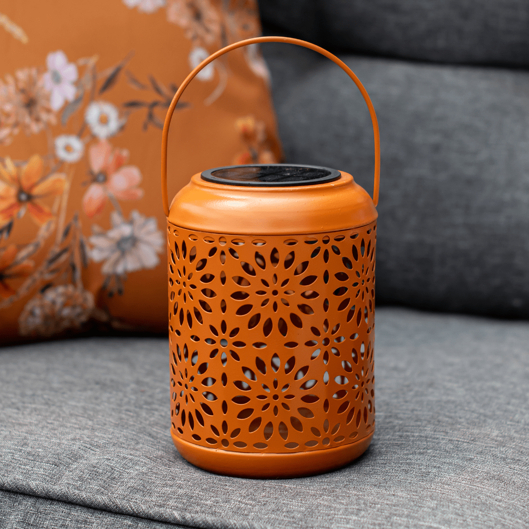 Orange metal lantern with floral cutouts on gray sofa, floral cushion in the background. Perfect for outdoor decor.