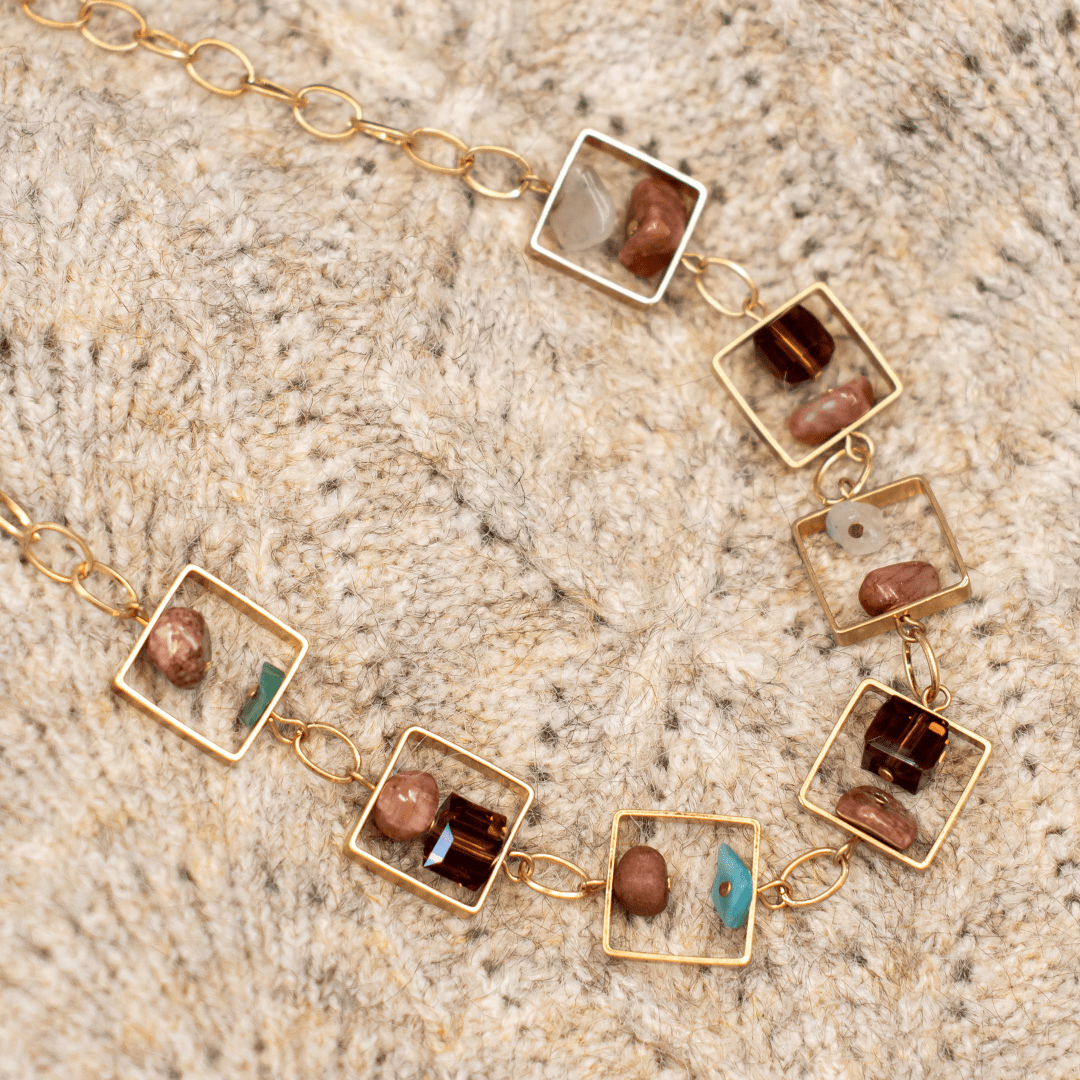 Gold chain necklace with square gemstone pendants on a knitted beige surface. Stylish boho-chic jewelry accessory.