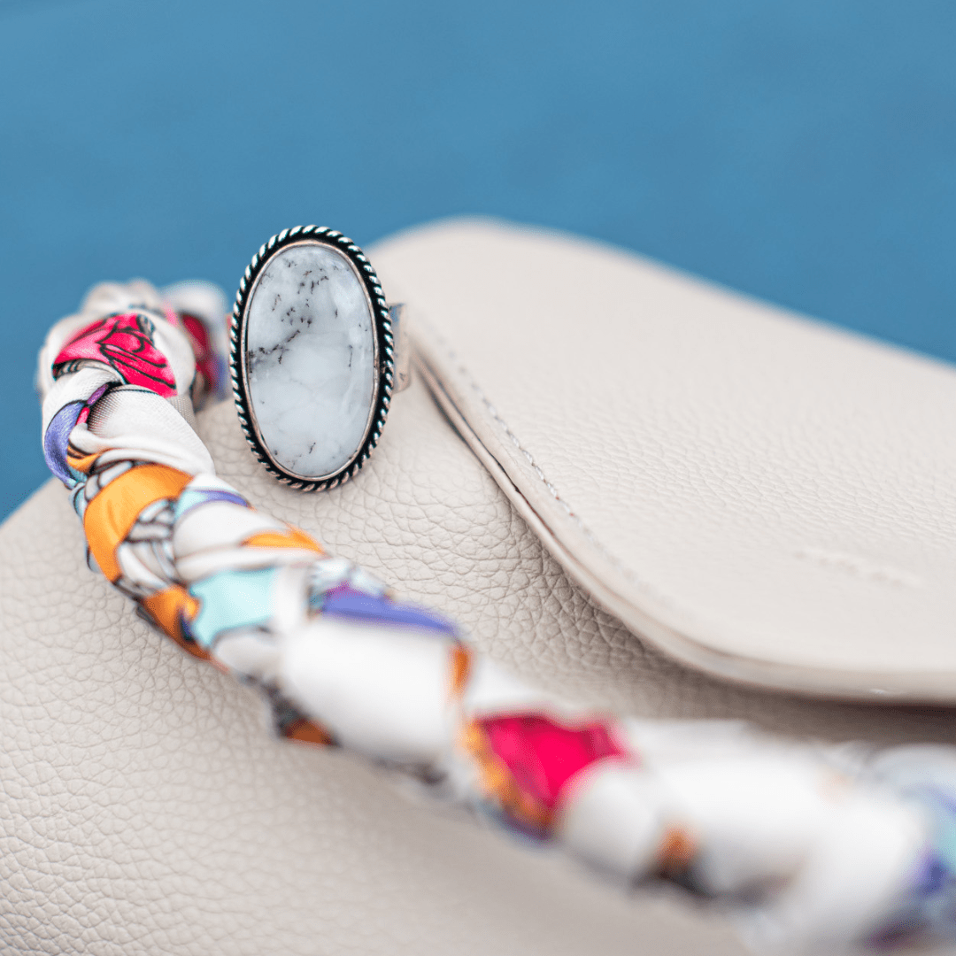 Oval marble ring on a beige leather handbag with colorful braided strap, set against a blue background.