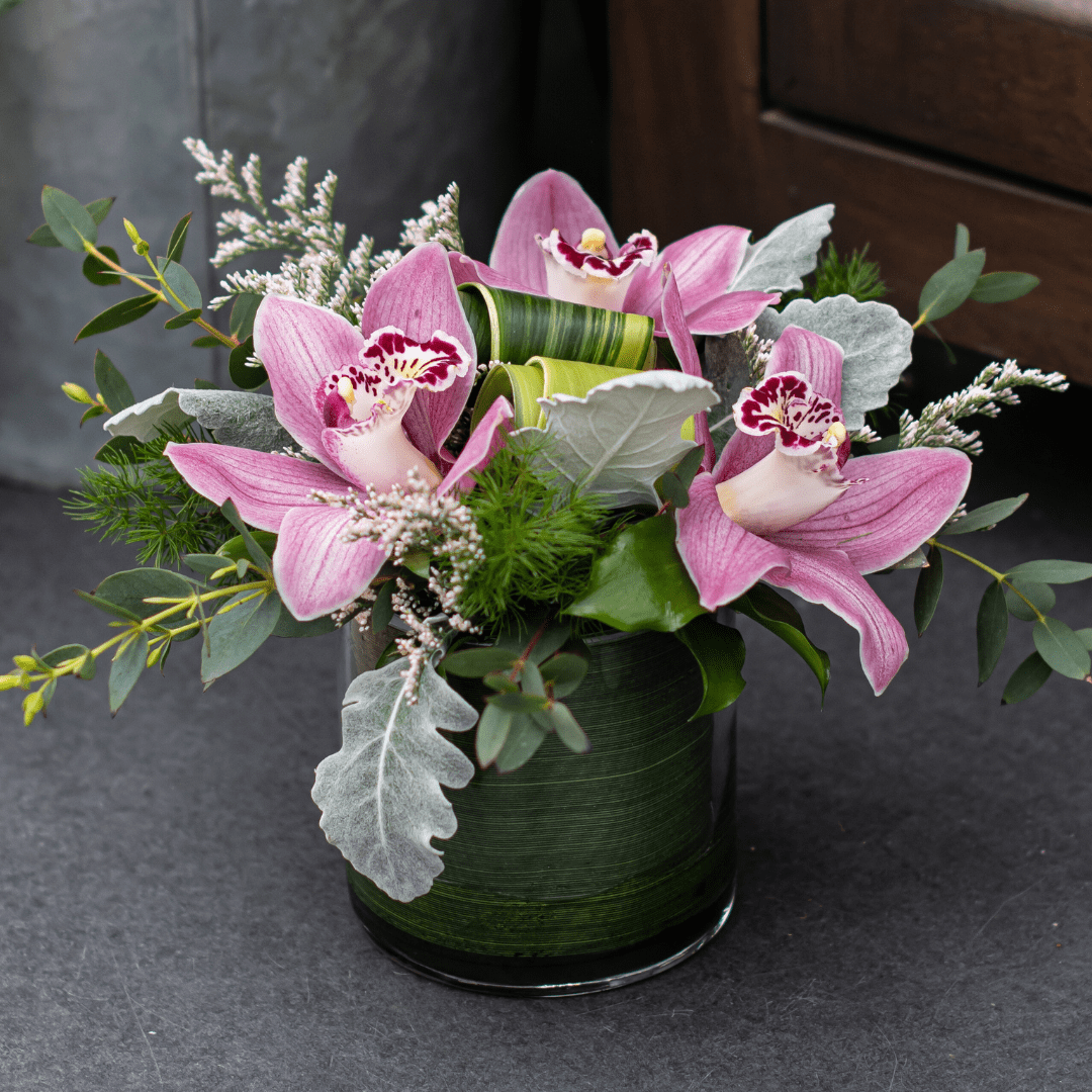 Pink orchids in a glass vase with lush green and silver foliage, creating a fresh and elegant floral arrangement.