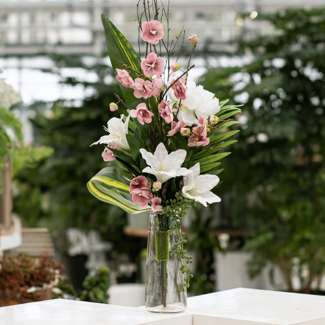 180 Elegant floral arrangement with pink and white flowers in a glass vase, set against a lush green backdrop.