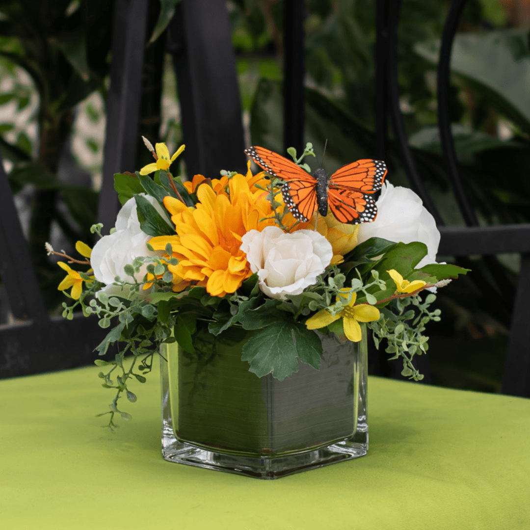 185 Vibrant flower arrangement with butterfly atop, featuring yellow and white blooms in a glass vase on a green surface.