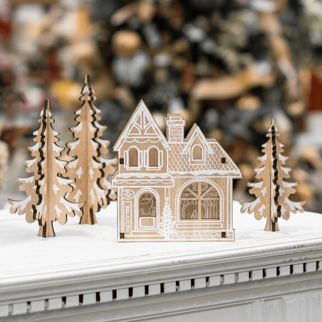 Decorative wooden Christmas village house with trees on a white table, featuring intricate festive details.