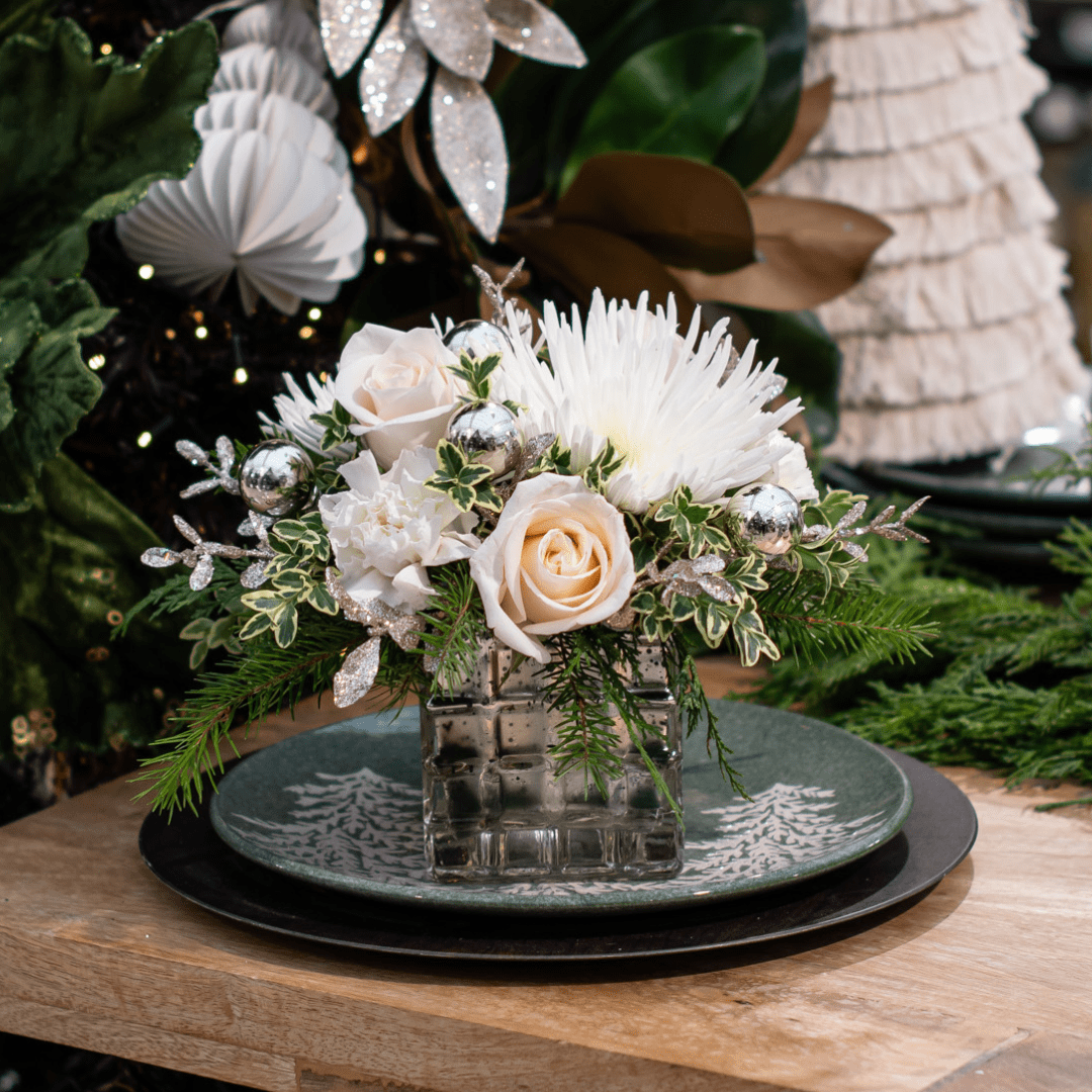 Elegant floral arrangement with white roses, greenery, and silver accents on a decorative plate. Perfect for holidays.