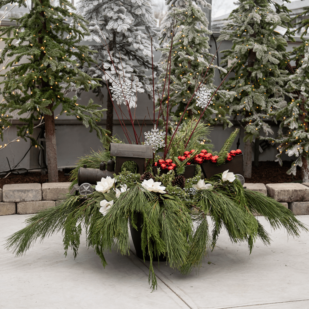 Design of the Day 2022 (5) Festive holiday planter with evergreen branches, snowflakes, and red accents, surrounded by snow-dusted Christmas trees.
