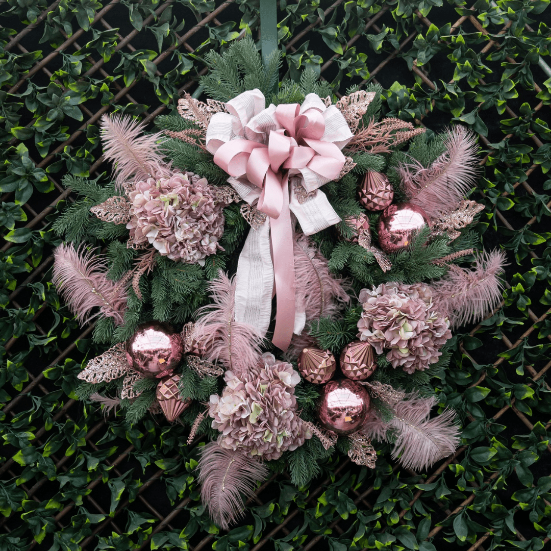 Design of the Day 2022 (8) Elegant pink holiday wreath with hydrangeas, feathers, ornaments, and a large bow against a green leafy background.