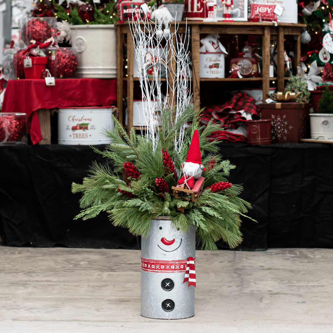 Mum Pot Email 2022-2023 - Slideshow (1) Festive Christmas arrangement in a smiling snowman pot with pine branches and gnome, surrounded by holiday decor.