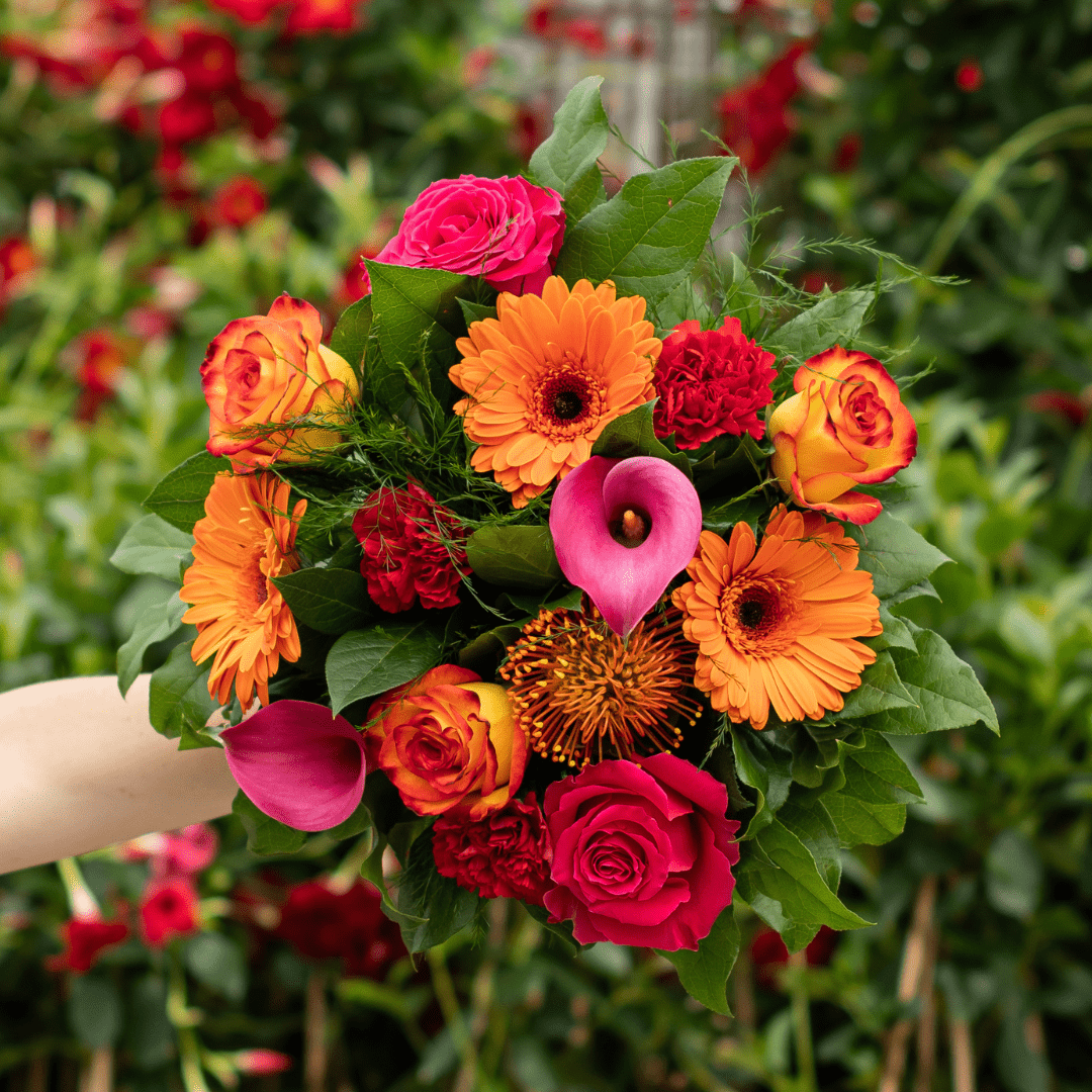 Vibrant bouquet with orange gerberas, pink calla lilies, and red roses against a lush green background.