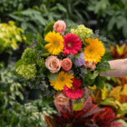 Vibrant floral bouquet featuring yellow daisies, pink roses, and greenery against a leafy garden background.
