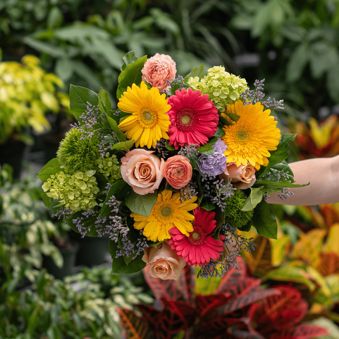 Vibrant floral bouquet featuring yellow daisies, pink roses, and greenery against a leafy garden background.