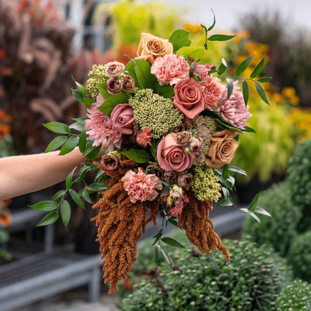 Hand holding vibrant bouquet with pink roses and various flowers, lush green leaves, garden backdrop.