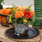 Festive autumn floral arrangement with roses, gerbera daisies, and a mini pumpkin on a rustic table setting.