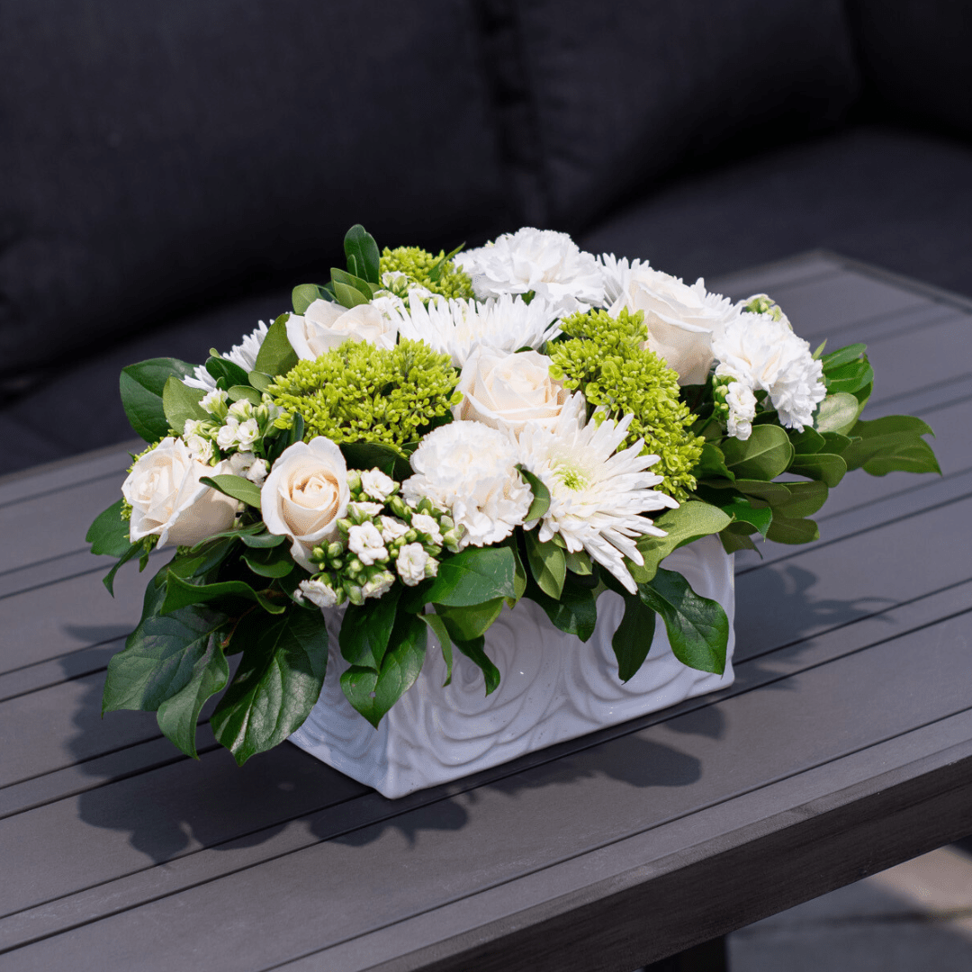 Elegant floral arrangement with white roses, chrysanthemums, and green accents in a rectangular white vase on a table.
