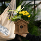 Decorative wooden birdhouse with yellow flowers and a bird figurine, held in a lush garden setting.