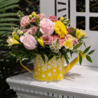 Bouquet of pink roses, yellow daisies, and greenery in a yellow floral watering can vase on a white table.