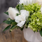 Elegant floral bouquet with white roses, carnations, and green hydrangeas in a white vase on a wooden table.