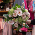 Bouquet of pink roses and white gerberas with greenery, held in a colorful shop setting.