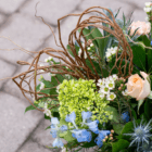 Elegant floral arrangement with twisted branches, green, white, and blue flowers on a textured stone background.