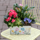 Colorful flower arrangement in a decorative planter on a yellow table, featuring pink and purple blooms with lush greenery.