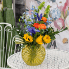 Vibrant flower bouquet in yellow glass vase on white table with floral backdrop and cactus decor.
