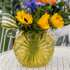Colorful bouquet in a yellow glass vase on a patterned table, featuring sunflowers and lush greenery.