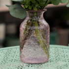 Ornate pink glass vase with floral designs sits on a decorative green table, holding a lush green plant.