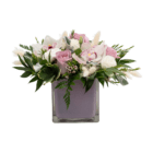 A floral arrangement with white phalaenopsis orchids, light pink-purple roses, white lisianthus, and white fluffy bunny tails arranged in a mauve-coloured glass cube.