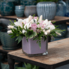 A floral arrangement with white phalaenopsis orchids, light pink-purple roses, white lisianthus, and white fluffy bunny tails arranged in a mauve-coloured glass cube. It sits on a wooden cart with blue pots blurred in the background.
