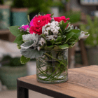 A floral arrangement in a clear glass vase lined with green bear grass that includes bright pink roses with faux diamonds in the middle, velvety dusty miller, white statice, and a fuchsia-coloured decorative butterfly pick. It sits on a wooden table.
