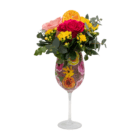 A floral arrangement with a bright, light pink, and yellow rose, as well as yellow kalanchoes and a faux orange slice arranged in a wine glass with a watercolour floral design on it.