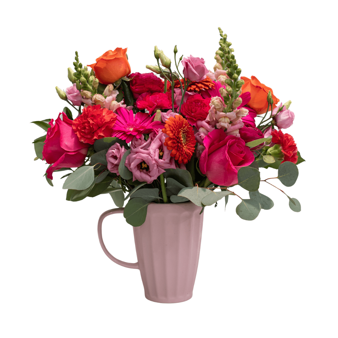 A fresh floral arrangement in a pink ceramic jug with bold pink and orange cut flowers, like gerberas, roses, snapdragons, carnations, and lisianthus.