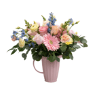 A fresh floral arrangement in a pink ceramic jug with light pink, yellow, peach, and blue coloured flowers, like gerberas, roses, delphinium, carnations, and snapdragons.