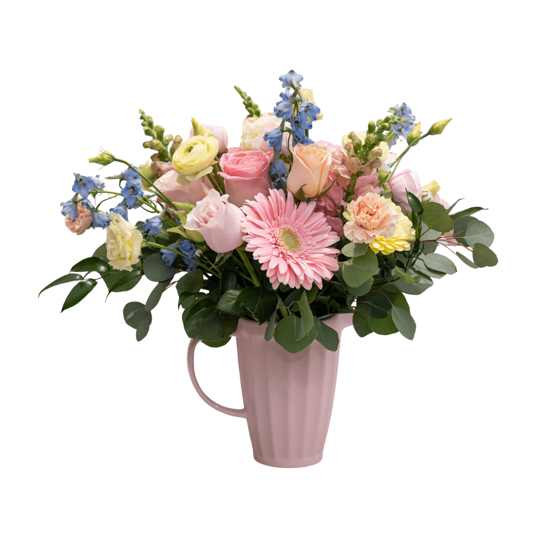 A fresh floral arrangement in a pink ceramic jug with light pink, yellow, peach, and blue coloured flowers, like gerberas, roses, delphinium, carnations, and snapdragons.