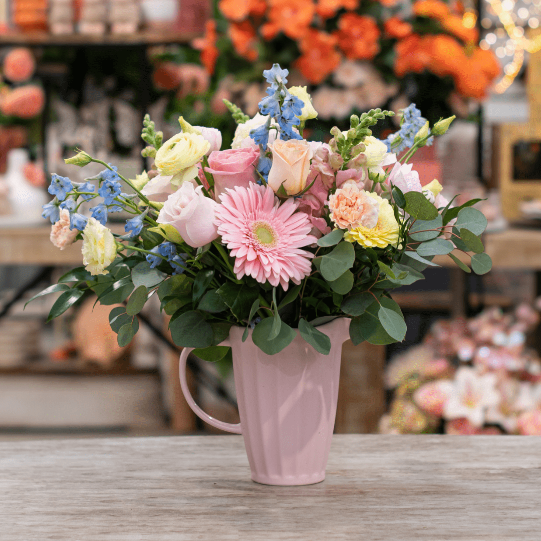 A fresh floral arrangement in a pink ceramic jug with light pink, yellow, peach, and blue coloured flowers, like gerberas, roses, delphinium, carnations, and snapdragons. It sits on top of a beige table.