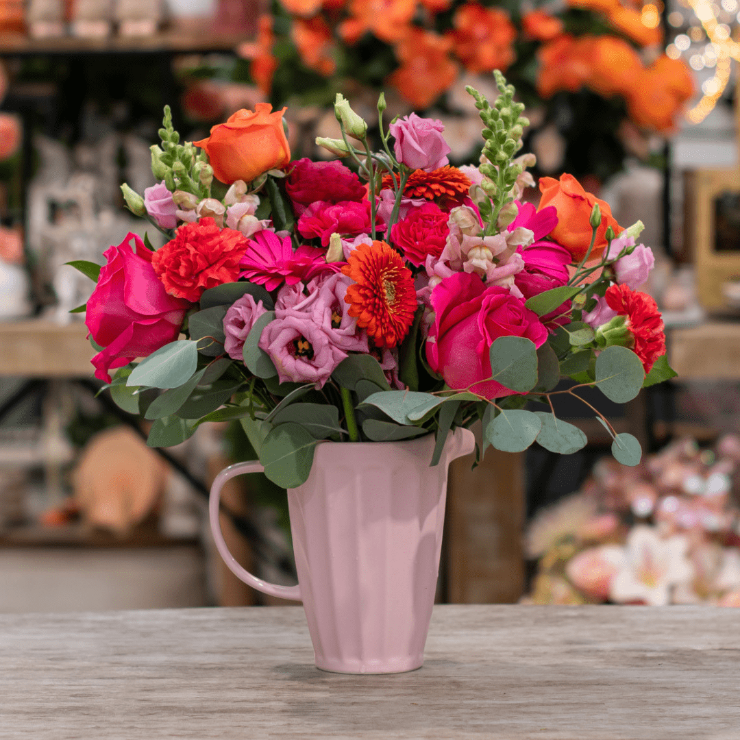 A fresh floral arrangement in a pink ceramic jug with bold pink and orange cut flowers, like gerberas, roses, snapdragons, carnations, and lisianthus. It sits on top of a beige table.