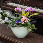 An indoor mixed planter in a sage container with a pink/purple Phalaenopsis orchid, pink kalanchoe, purple African violet, and faux stems of lavender. The planter sits on a wooden piano bench.