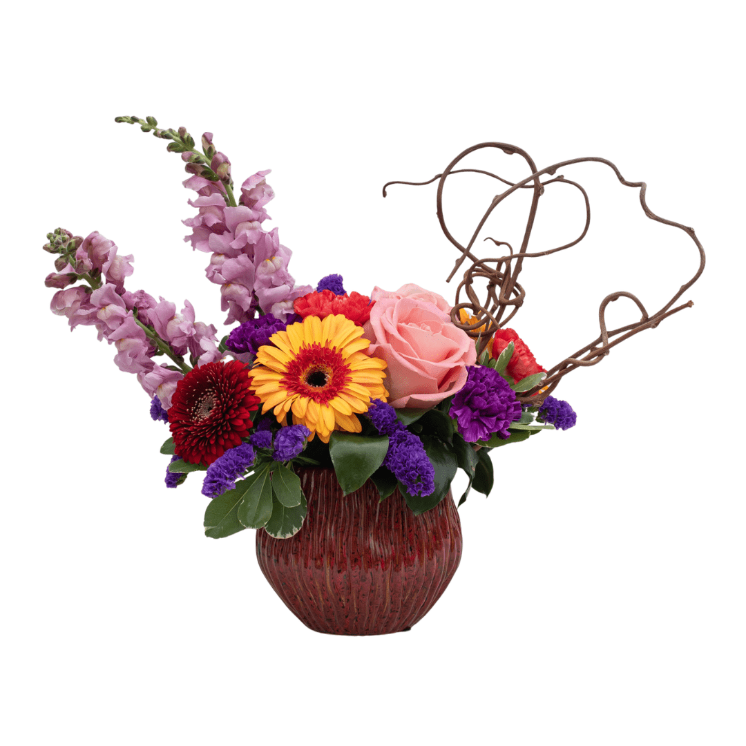 298 An image of a floral arrangement in a red ceramic container with pink snapdragons, coral carnations, red and yellow/orange gerberas, purple statice, and whimsical decorative branches.