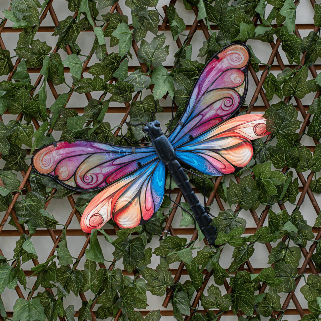 An image of a pink, purple, and blue metal dragonfly on a wall of artificial greenery.