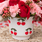A white ceramic pot with red and green cherries on it sits on a light wicker place setting and has red and pink flowers arranged in it.