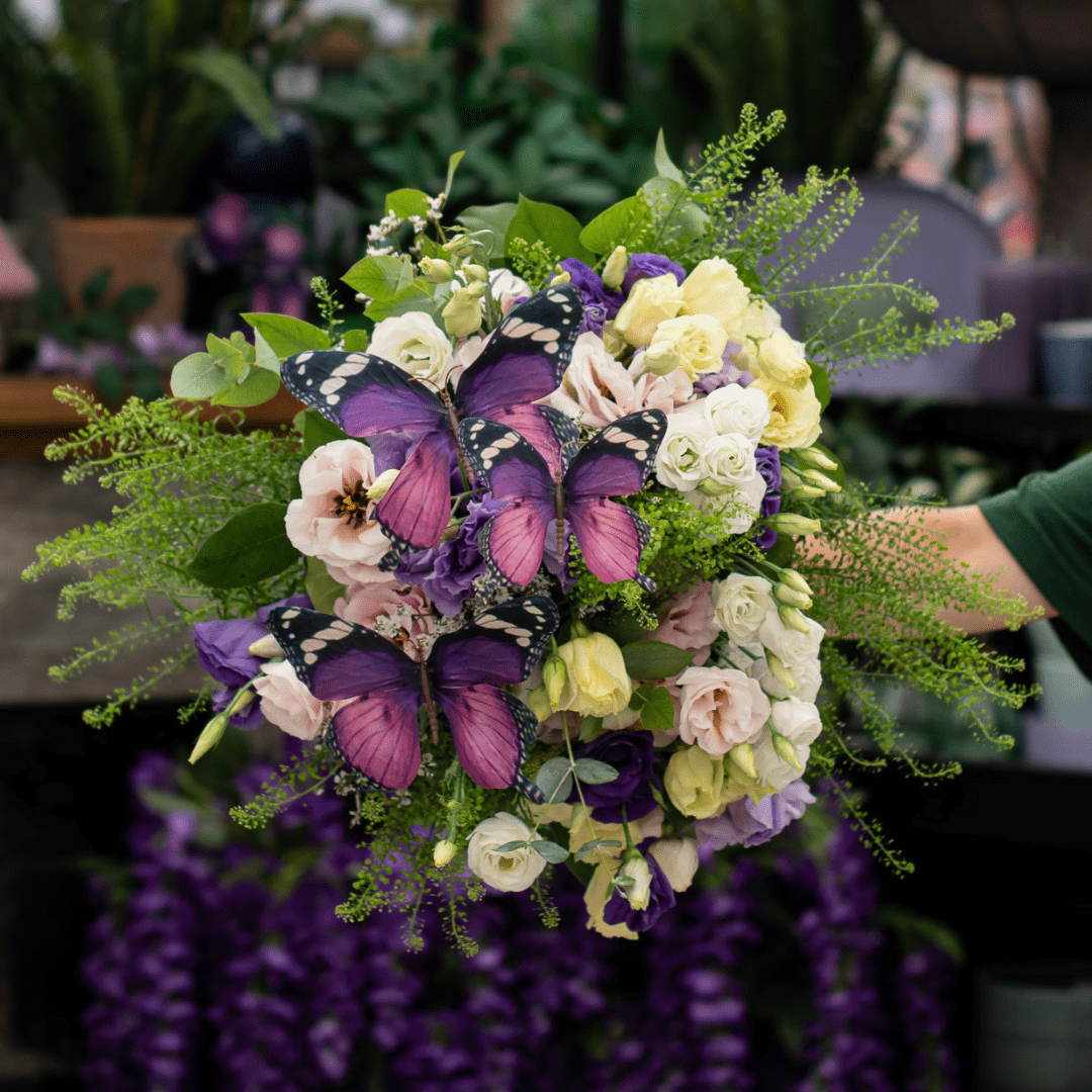A fresh floral bouquet with pale pink and yellow lisianthus, as well as white and purple, with eucalyptus and other greenery accents, and faux purple butterflies floating over the foliage and blooms.
