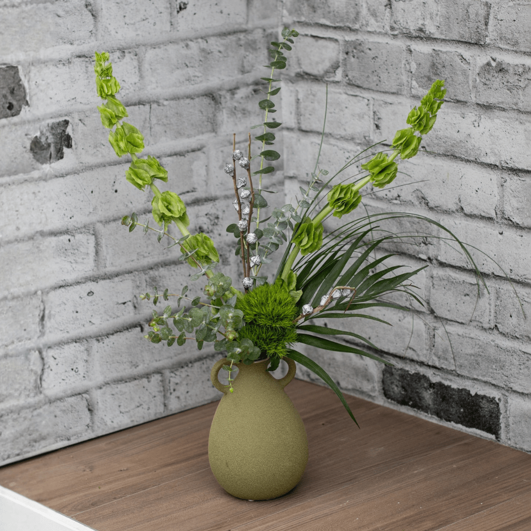 Fresh greenery and dried eucalyptus pods in an olive-coloured textured vase with two small decorative handles on the side. It sits on dark wood flooring with white and grey bricks behind it.