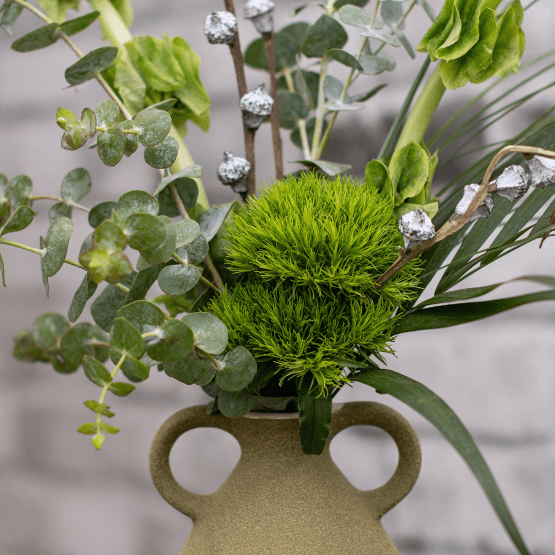 A close-up of fresh greenery in an olive-coloured textured vase with two small decorative handles on the side.